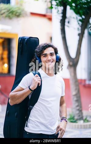 Portrait of confident young man carrying guitar in bag while standing on street, Santa Cruz, Seville, Spain Stock Photo