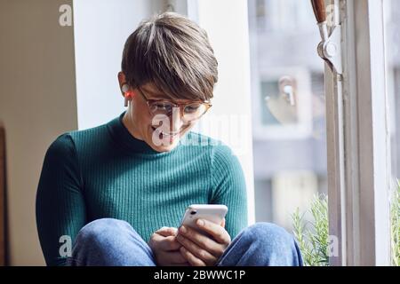 Happy woman with smartphone and earbuds at the window at home Stock Photo