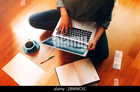 From above anonymous female freelancer sitting on floor near cup of coffee and open planner and browsing laptop while working on design project at hom Stock Photo