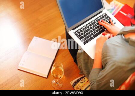 From above anonymous woman sitting on floor near glass of wine and open planner and browsing laptop while working on remote project at home Stock Photo