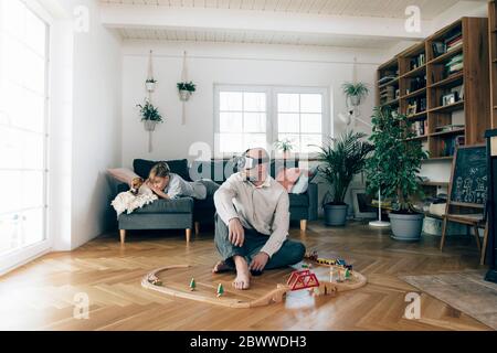 Father sitting in the middle of toys, using VR glasses, while son is lying on couch Stock Photo