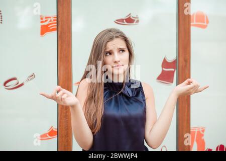 It is confusing. Portrait dumb looking woman arms out shrugs shoulders I don't know what to buy, store window wall on background. Negative human emoti Stock Photo