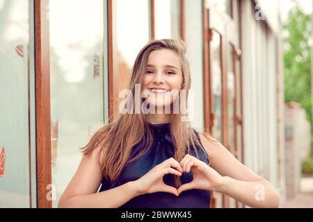 Young woman outdoors in European city with beautiful beaming smile lit by warm glow of sun shining down on summer day. Girl with hands shaped like hea Stock Photo