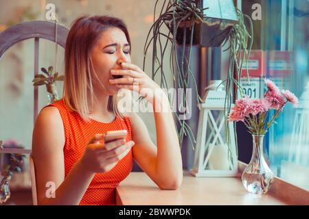 Tired woman yawning while is working on the phone in a restaurant coffee shop. Sleepy and bored by the message she received in her home kitchen, near Stock Photo