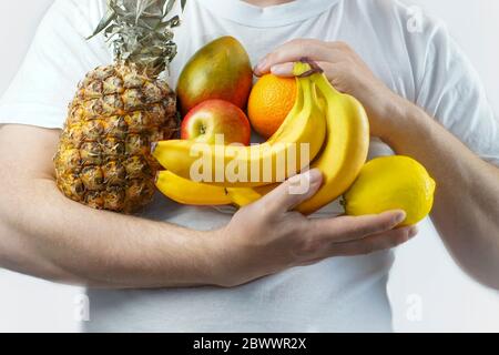 Cook man in a white T-shirt holds fruits in his hands: pineapple, bananas, lemon, orange, apple, mango Stock Photo