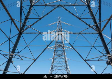A high voltage electricity pylon seen through a close up of the lattice structure of a second tower with a blue sky background Stock Photo