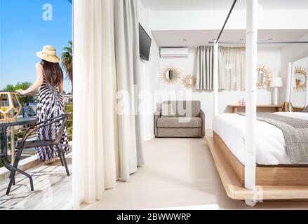 Young woman in romantic style dress standing on balcony of modern resort hotel bedroom. Light open space room interior with white, gray, wooden furnit Stock Photo