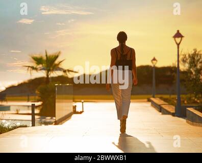 Back view of female silhouette going on evening sunset background of resort hotel. Woman walking by tropic street way with palms and mountain Stock Photo