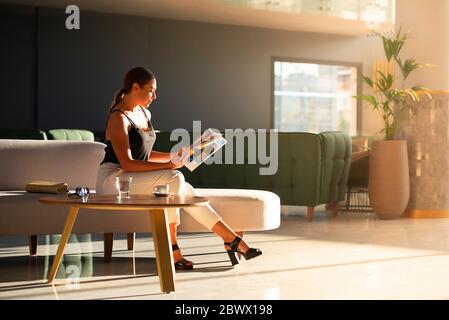 Young adult woman reading magazine on sofa in light modern interior of hotel reception, office building hall, waiting area background Stock Photo