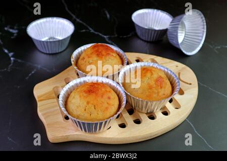 Homemade Banana Muffins in Mold on Wooden Bread Board with Empty Aluminum Muffin Mold on Kitchen's Black Marble Table Stock Photo