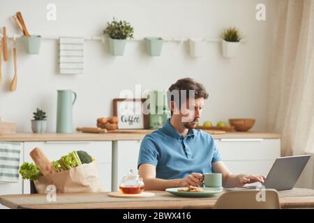 Young man sitting at the table in the kitchen drinking coffee and working online using his laptop Stock Photo