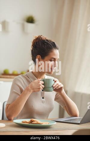 Young woman sitting at the table and drinking coffee while using laptop at home Stock Photo