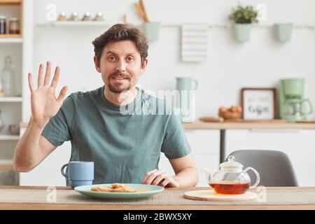 Portrait of young man looking at camera and waving his hand while having breakfast in the kitchen Stock Photo