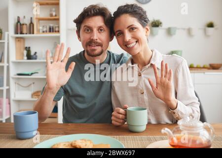 Portrait of young family smiling at camera and waving while sitting at the table and having breakfast in the kitchen Stock Photo