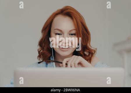 Pretty positive redhead woman gives online consultation, sits in front of opened laptop computer, has discussion with corporate client, smiles happily Stock Photo