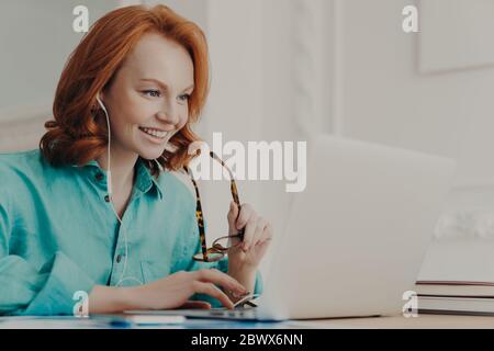 Side view of happy lady freelancer communicates with client via video computer call, uses laptop computer and earphones, types information. Woman tuto Stock Photo