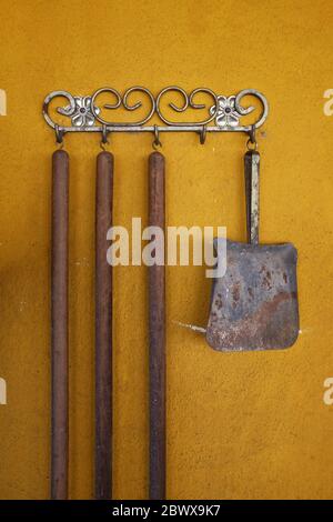 Metallic hand shovel hanging on a yellow wall next to three wooden sweeper handles Stock Photo