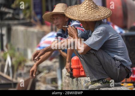 Yangshuo, China - August 2019 : Two Chinese men wearing traditional wicker conical hats smoking cigarettes and chatting during work break Stock Photo