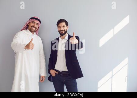 Arab and european businessmen show thumb up stand smiling at the gray background. Stock Photo