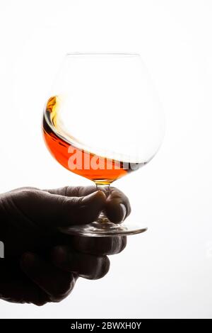 Man's hand holding snifter with cognac. Splash of a drink in a glass. White bright background. Stock Photo