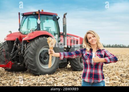 A young woman in a field on a background of a tractor with spike of wheat in her hands. Stock Photo
