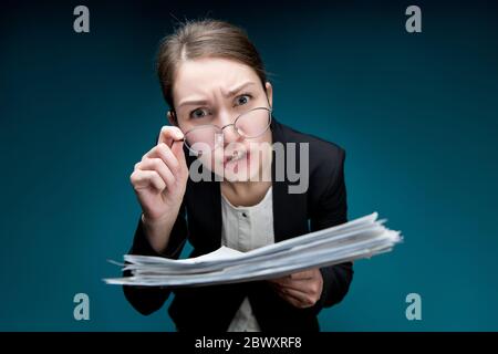 A young woman puts glasses on her nose and looks in camera with angry surprise. Director dissatisfied with paperwork or errors in them. Teacher scream Stock Photo