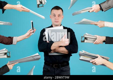 A tired office employee in a black shirt and folders with documents in his hands looks into the void. At this time, many hands pull folders, documents Stock Photo