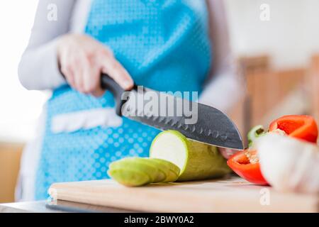 Close up of woman in apron slicing zucchini on cutting board in kitchen. Healthy meal preparation concept. Stock Photo