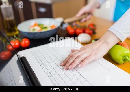Close up of female hand on laptop keyboard while holding cooking pan with vegetables. Online recipe concept. Stock Photo