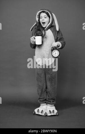 Drinking coffee energy. Morning routines. Adorable bunny hold alarm clock. Small girl in bunny costume. Child rabbit kigurumi. Girl bunny pajamas. Bunny kid red background. Baby animal. Counting time. Stock Photo