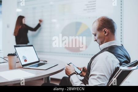 A man entrepreneur sitting at the conference table in a boardroom and reading messages on his cellphone while his female colleague using an electronic Stock Photo