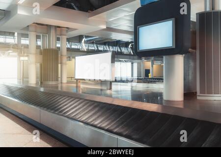 A baggage claim area in a hall of a modern airport arrival zone with conveyor belt and a mock-up of white empty advertising or information billboard o Stock Photo