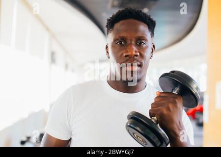 Young African American man standing and lifting a dumbbell with the rack at gym. Male weight training person doing a biceps curl in fitness center Stock Photo