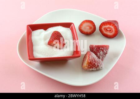 Fresh raw cut strawberries and sugar in red sugar bowl on white plate on pink background Stock Photo
