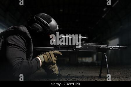 Photo of a soldier in helmet and ammunition laying and aiming sniper rifle profile view on dark background. Stock Photo