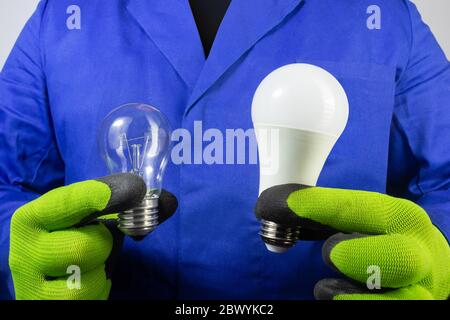 Photo of electrician in blue robe and gloves holding old bilb and new led light bulb close-up view. Stock Photo