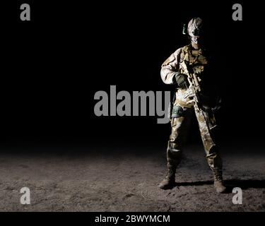 Photo of a fully equipped soldier standing with automatic rifle with scope on a sand desert ground on dark background full body front view. Stock Photo