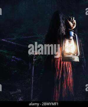 Photo of a female witch holding lantern and cane in dark woods gloomy, scary background. Stock Photo