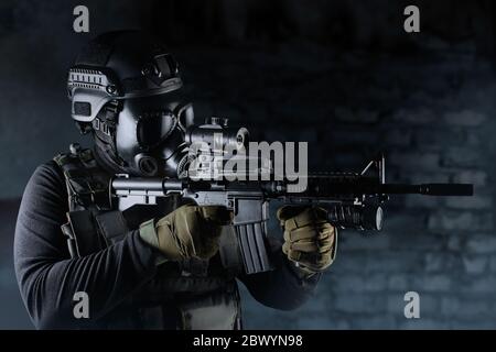 Photo of a fully equipped soldier in black armor tactical vest, gas mask, automatic rifle, gloves and helmet aiming profile view on black brick backgr Stock Photo