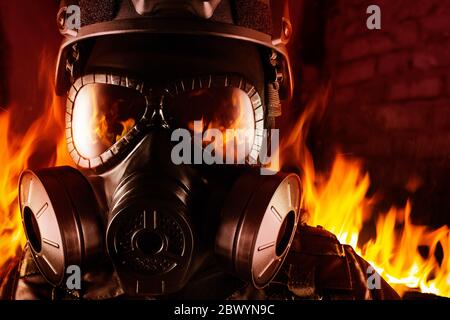 Photo of a fully equipped soldier in black armor tactical vest, gas mask standing in fire on brick background closeup front view. Stock Photo