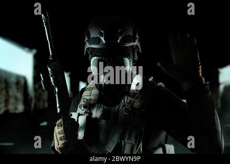 Photo of a fully equipped soldier in black armor tactical vest, gas mask, automatic rifle, gloves and helmet standing and showing tactical sign on bla Stock Photo