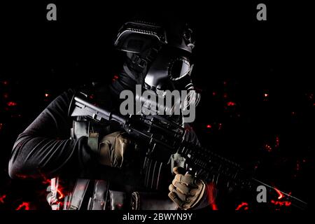 Photo of a fully equipped soldier in black armor tactical vest, gas mask, automatic rifle, gloves and helmet aiming profile view standing in ashes on Stock Photo