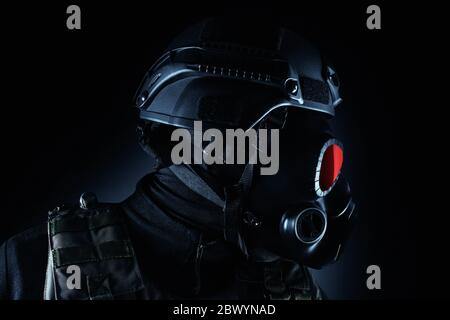 Photo of a fully equipped soldier in black armor tactical vest, gas mask with red eye and helmet standing on black background closeup profile view. Stock Photo