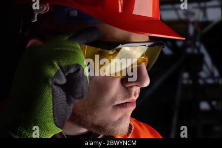 Photo of a worker in orange jacket outfit with protective gloves and helmet putting on glasses on construction area background. Stock Photo