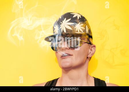 Girl in sunglasses and cap with leaves of marijuana smokes on yellow background Stock Photo