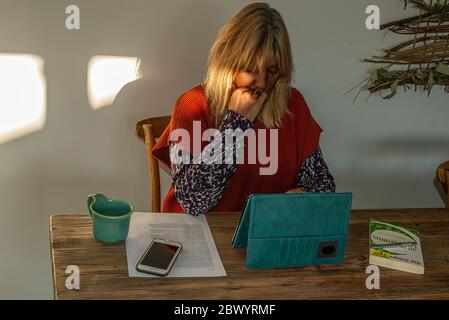 Woman during lockdown, wearin a red knitted top, using an iPad, with coffee mug, smart phone, paper and book. Dappled sunlight on the wall beside her. Stock Photo