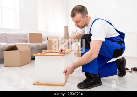 Repairman Assembling Furniture In Flat After Relocation, Wearing Blue Coverall Stock Photo
