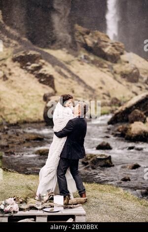 Destination Iceland wedding, near Kvernufoss waterfall. Wedding couple on the shore of a mountain river. The groom wears the bride in a woolen blanket Stock Photo