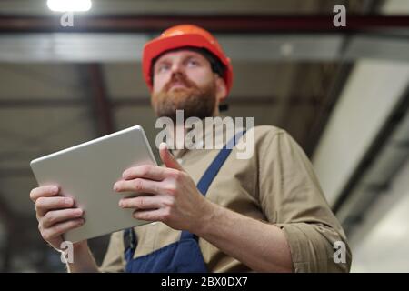 Below view of young bearded engineer in hardhat using tablet while controlling work at factory Stock Photo