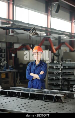Portrait of content attractive Asian metalworking specialist in hardhat standing at table with metal details in workshop Stock Photo